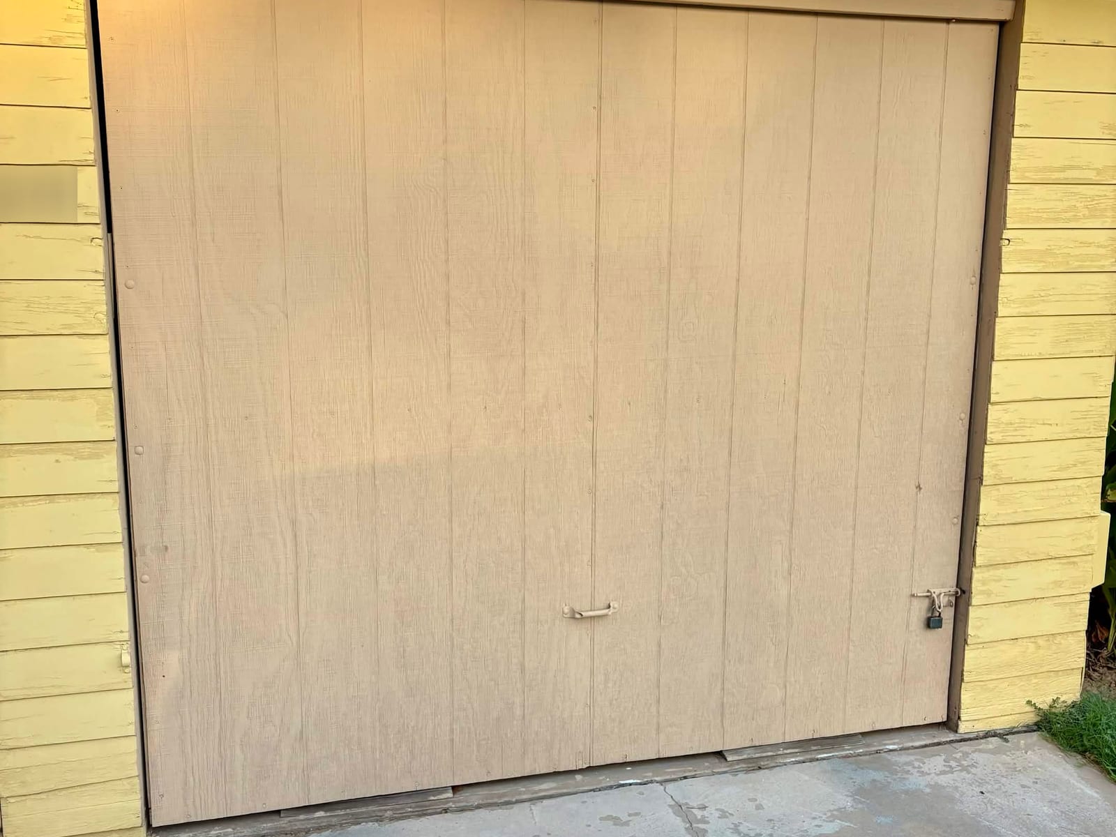 Before: old tan one-piece tilt-up garage door with a handle and padlock on a yellow-sided garage in Phoenix
